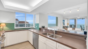 Een moderne keuken met witte kasten, een groot schiereiland met een spoelbak en uitzicht op zee door grote ramen.