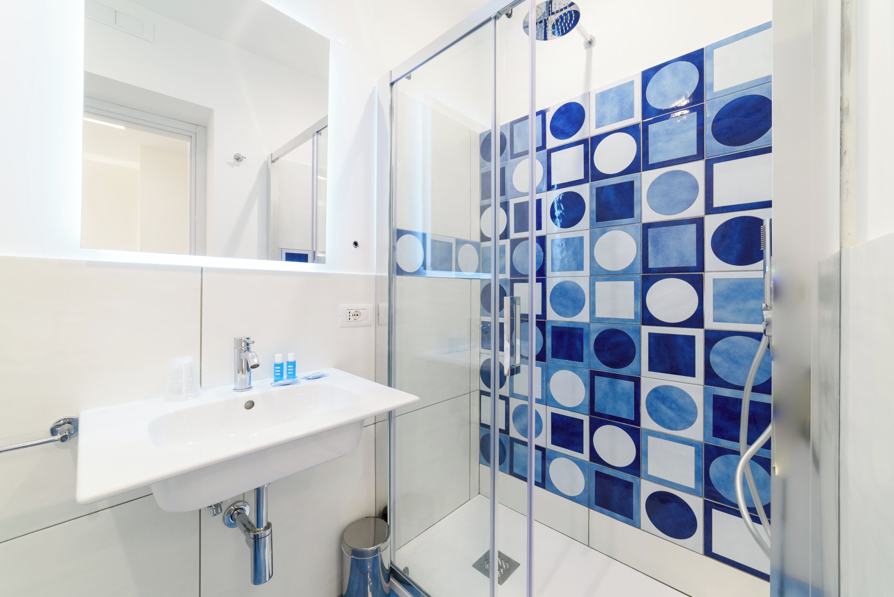 A modern bathroom with a white sink, glass shower enclosure, and blue geometric tile backsplash.