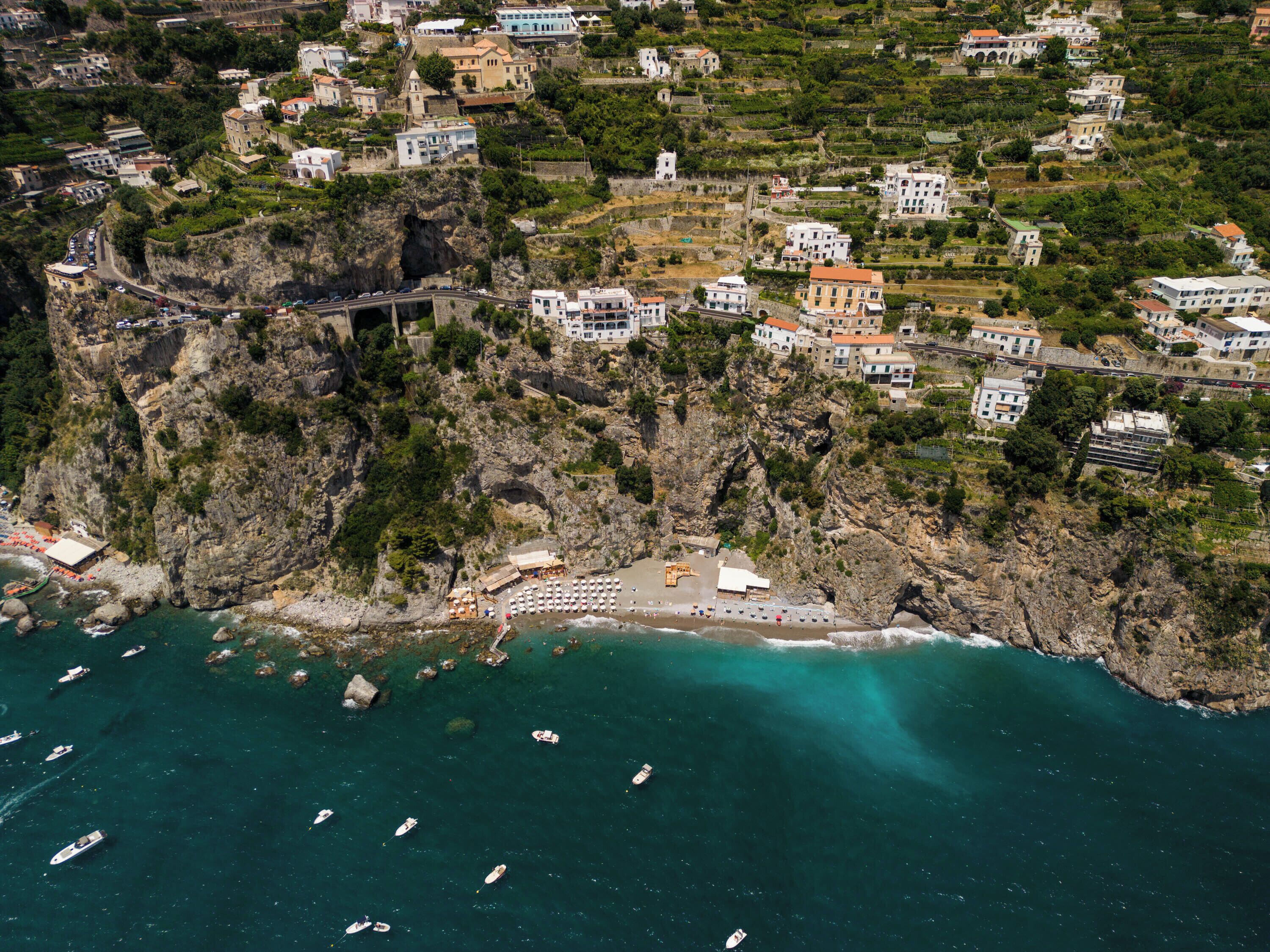 Vista aerea di una cittadina costiera con edifici su una collina, una spiaggia con barche e una strada che collega la zona.