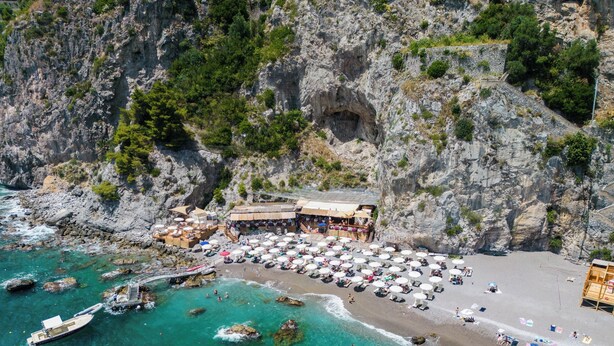 Vista aérea de una zona costera con playa, un acantilado rocoso y un edificio con terraza.