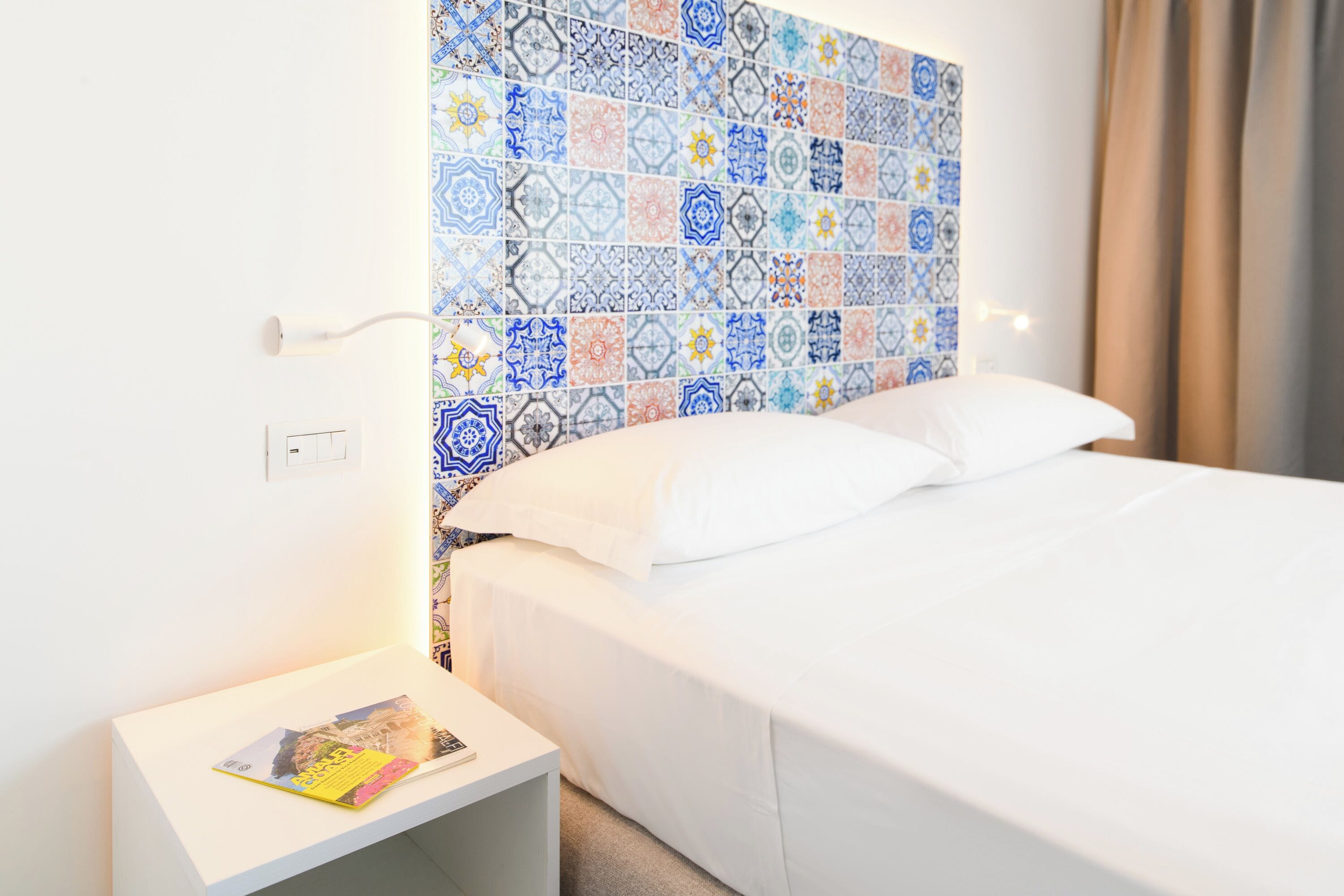 Una habitación de hotel con cabecera de cama de baldosas estampadas, una cama blanca y una mesita de noche con una revista.