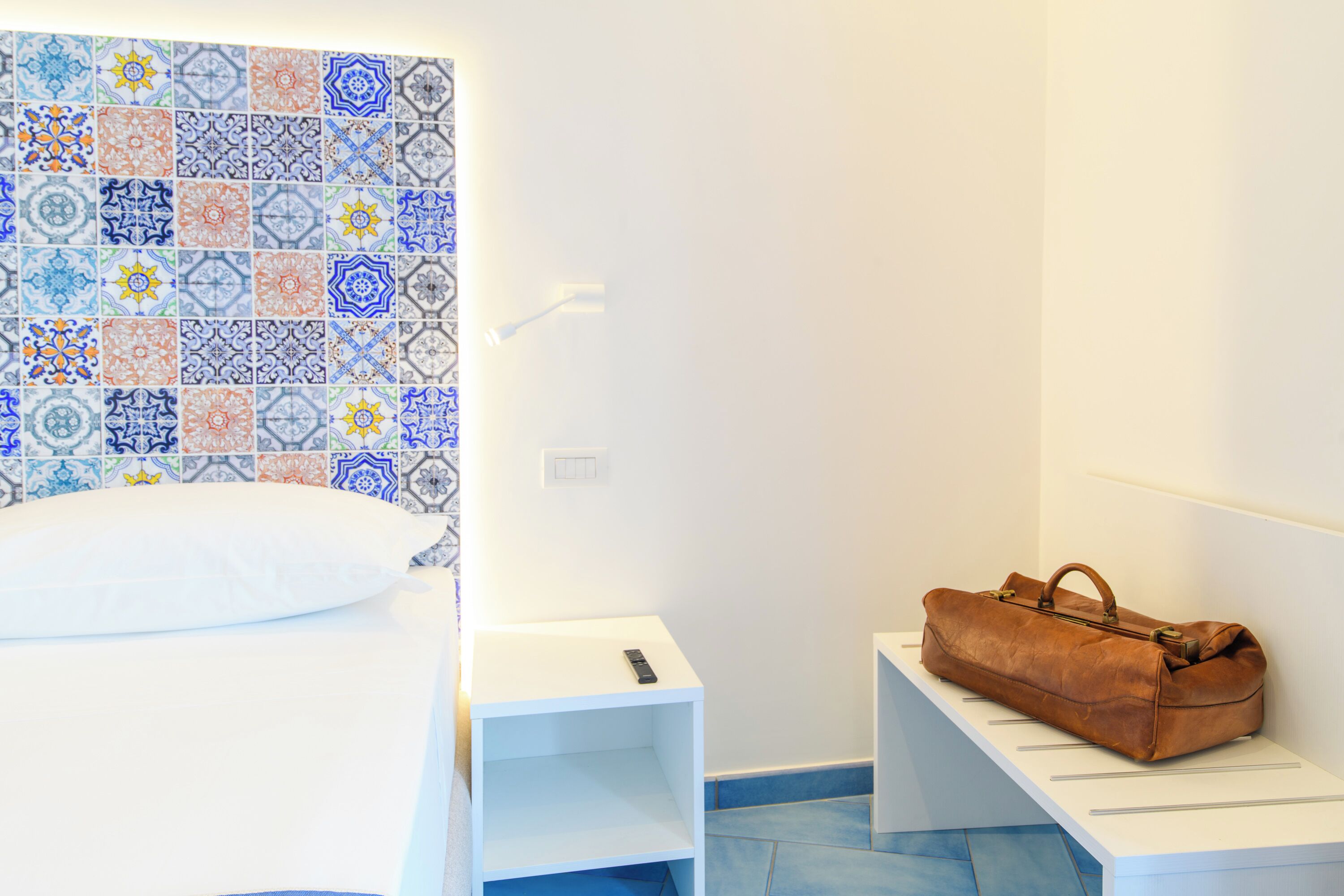 A neatly arranged hotel room with a bed, a small bedside table, a bench, and a wall with colorful tiles.