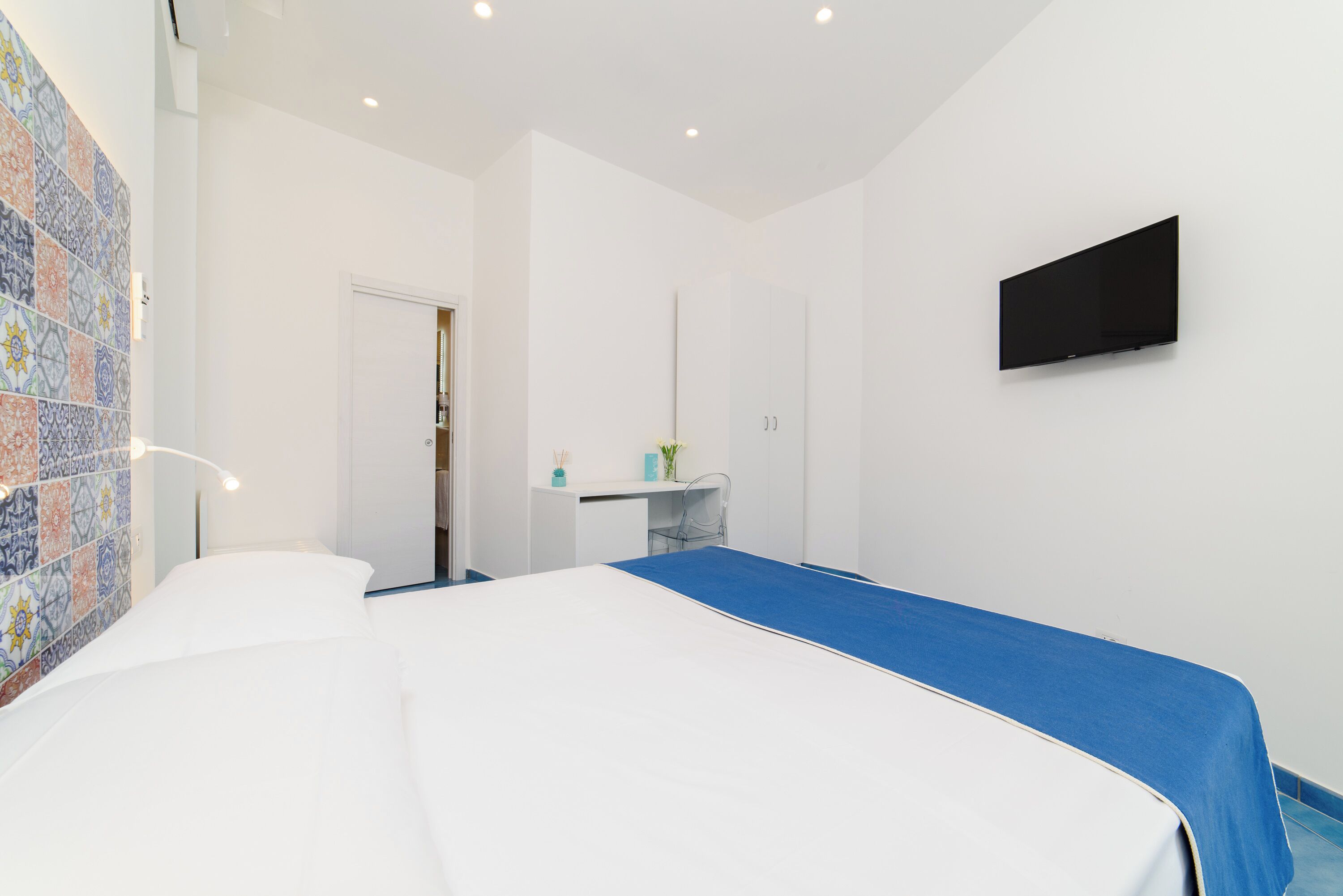 A bedroom with a bed, a desk, a chair, a TV, and a door.