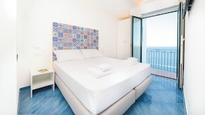 Un dormitorio moderno con una cama grande, una mesita de noche, una pared de azulejos coloridos y un balcón con vista al mar.