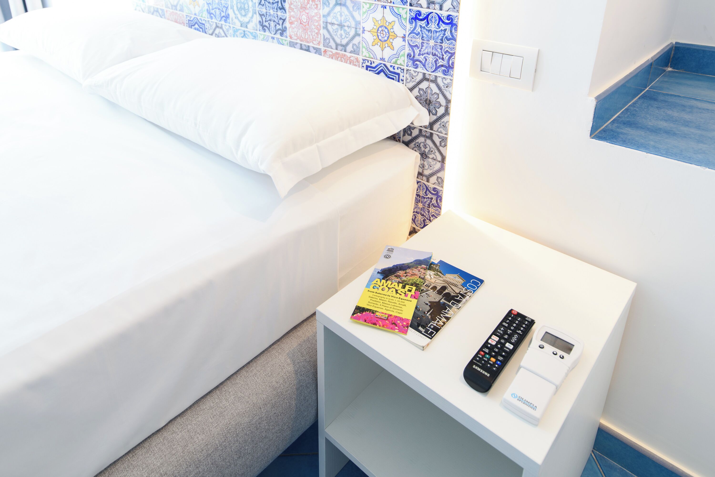 Una cama bien hecha con ropa de cama blanca, una mesita de noche pequeña con un control remoto, una revista y un dispositivo digital.