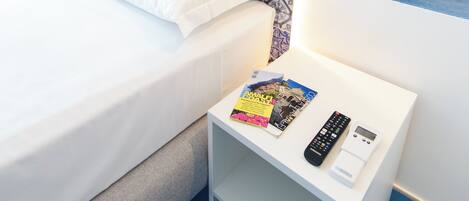 Una cama bien hecha con ropa de cama blanca, una mesita de noche pequeña con un control remoto, una revista y un dispositivo digital.