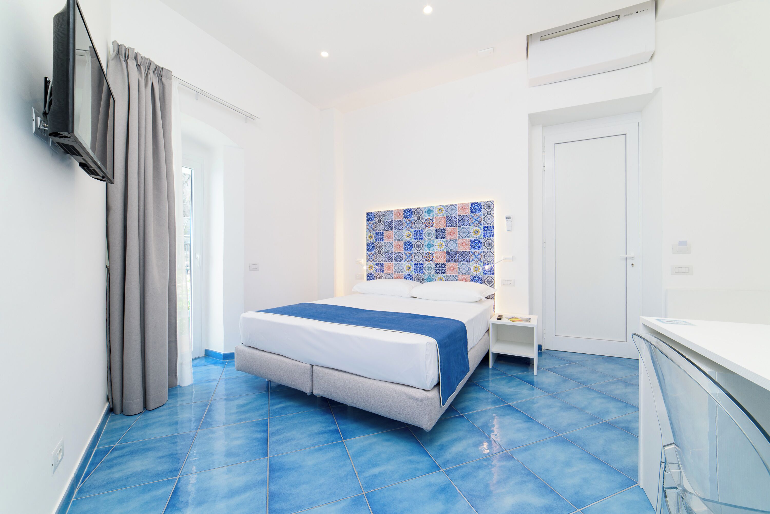 A modern hotel room with a bed, a nightstand, a TV, and a bathroom with a white door and a tiled backsplash.