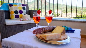 A table with a plate of cheese and cured meats, two glasses of red beverage, and a wicker sofa with a polka dot pillow in a balcony setting. - I Mandorli - Lovely loft in Sardinia (Loceri)