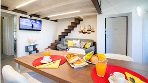 A modern living room with a wooden dining table set for breakfast, a staircase, and a TV mounted on the wall. - I Mandorli - Lovely loft in Sardinia (Loceri)