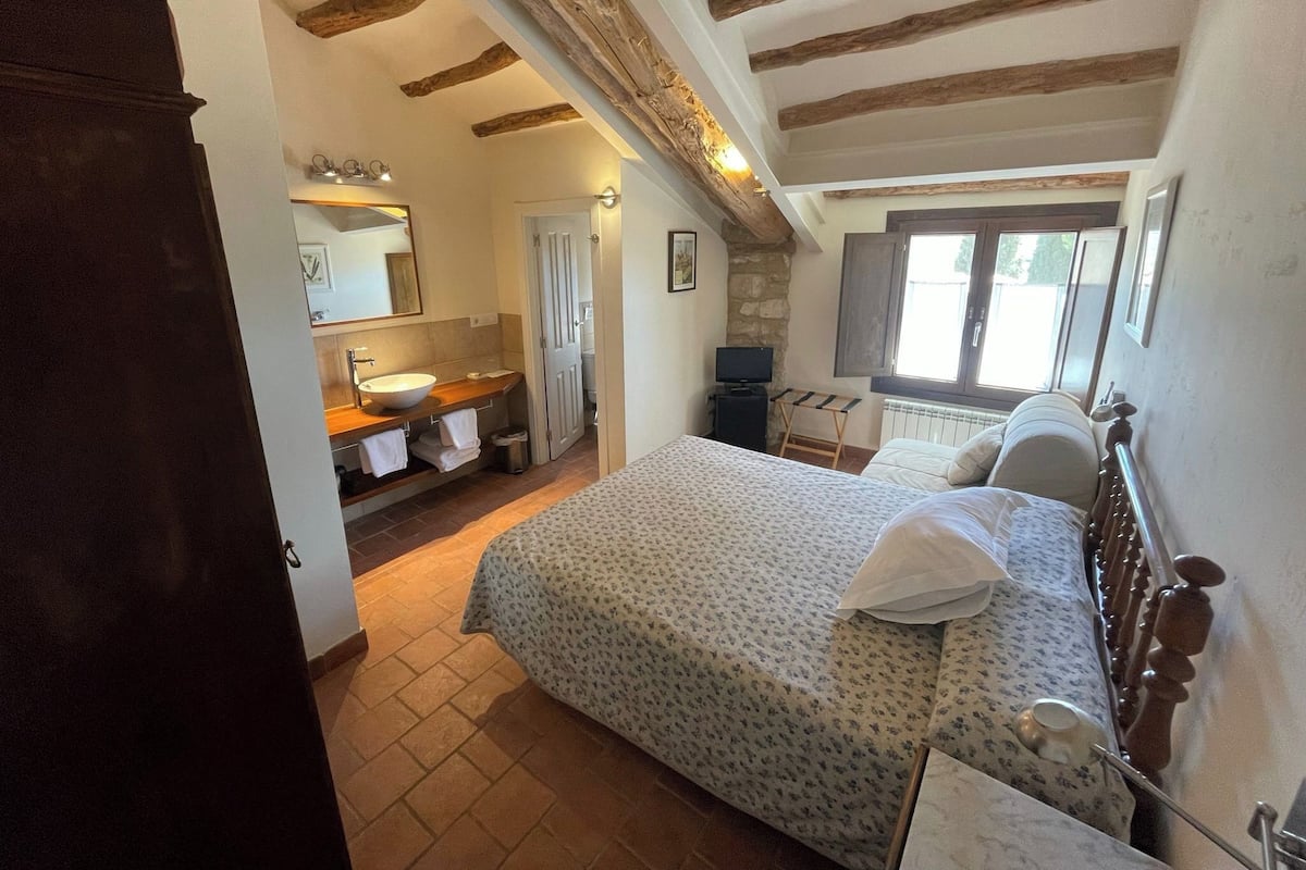 A bedroom with a bed, a wooden headboard, a window, a stone wall, and a bathroom with a sink and mirror.