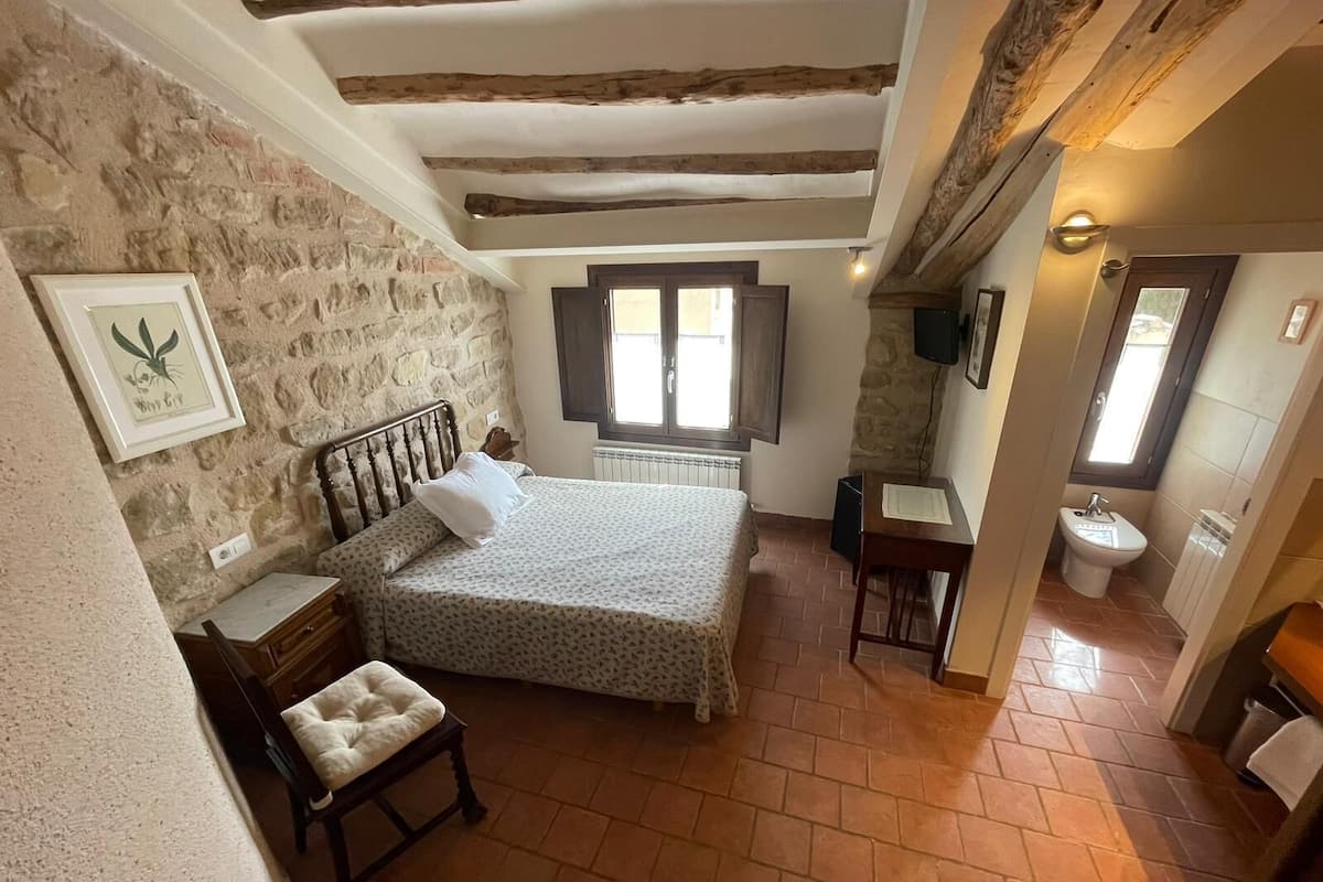 A bedroom with a stone wall, a bed with a headboard, a small table, a chair, a window, and a bathroom with a sink and toilet.