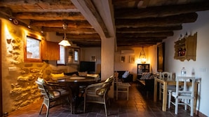 A rustic interior with a stone wall, wooden ceiling beams, a round dining table with wicker chairs, and a small kitchen area with a wooden table and chairs. - Torre alta de Ram (Alcaniz)