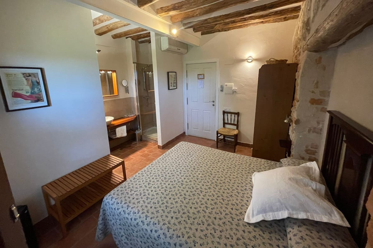 A bedroom with a bed, a wooden bench, a chair, a wardrobe, and a bathroom with a shower and sink.