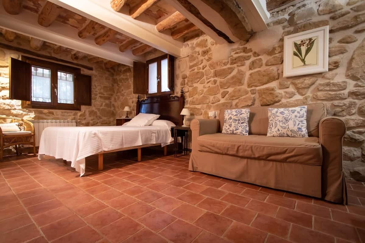 A rustic bedroom with a stone wall, a wooden bed, a brown sofa, and a small table.
