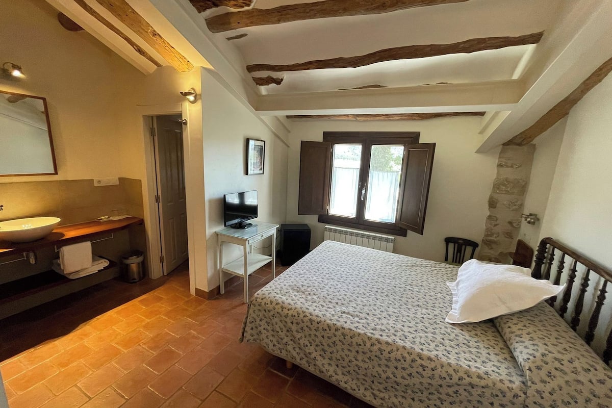 A bedroom with a bed, a desk, a television, a window, and a bathroom with a sink and mirror.