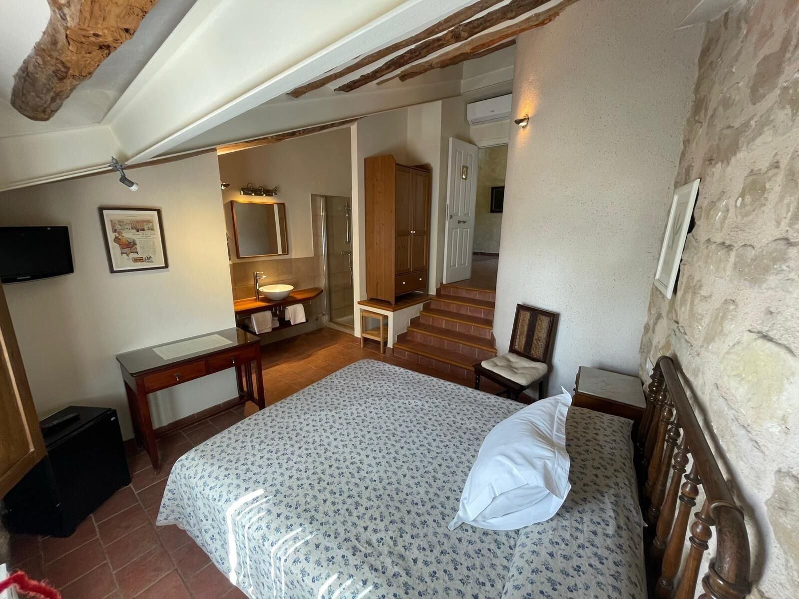 Un dormitorio con cama, cabecera de madera, mesita de noche, escritorio, silla, espejo, lavabo y ducha.