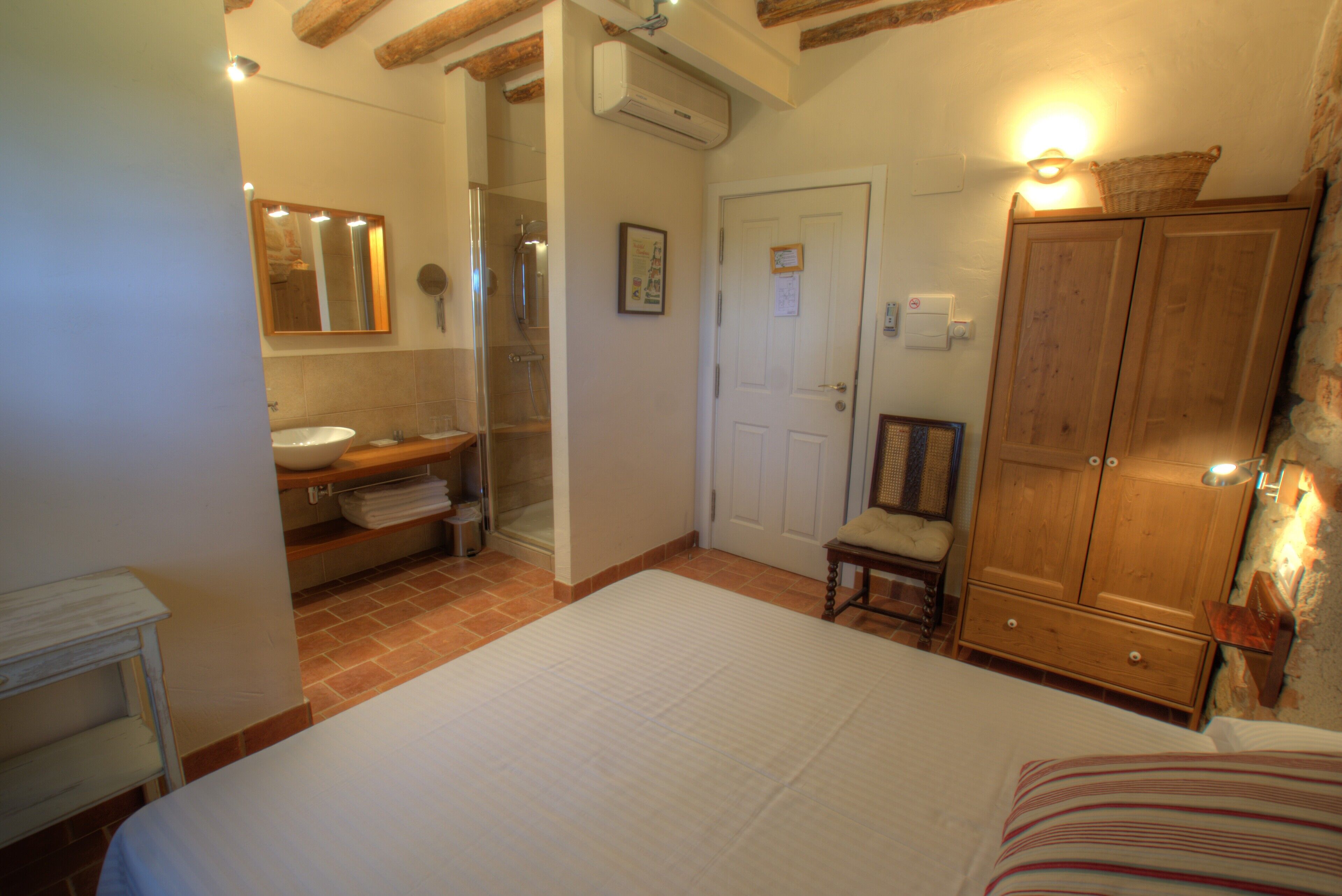 A bedroom with a bed, a wooden wardrobe, a bathroom with a sink and shower, and a chair.