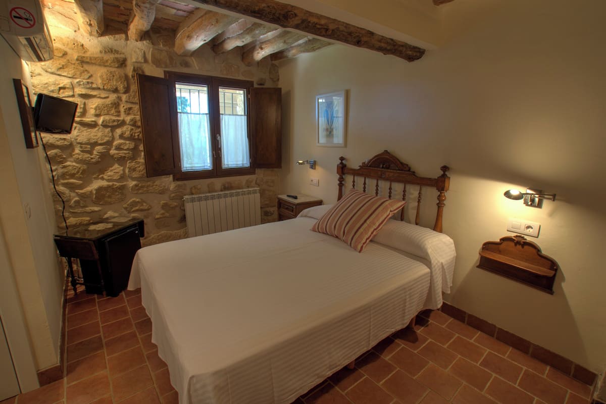 A bedroom with a stone wall, wooden ceiling beams, a large bed with a wooden headboard, a nightstand, a bedside table, and a window with curtains.