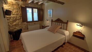 Un dormitorio con pared de piedra, vigas de madera en el techo, una cama grande con cabecera de madera, una mesita de noche y una ventana con cortinas.