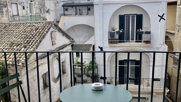 En balkon med et rundt bord og to polstrede stole, med udsigt over en gårdhave med bygninger.