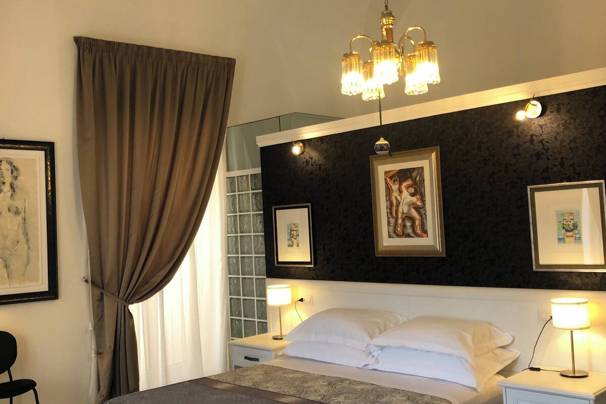 A hotel room with a bed, two bedside tables, a chandelier, and artwork on the walls.