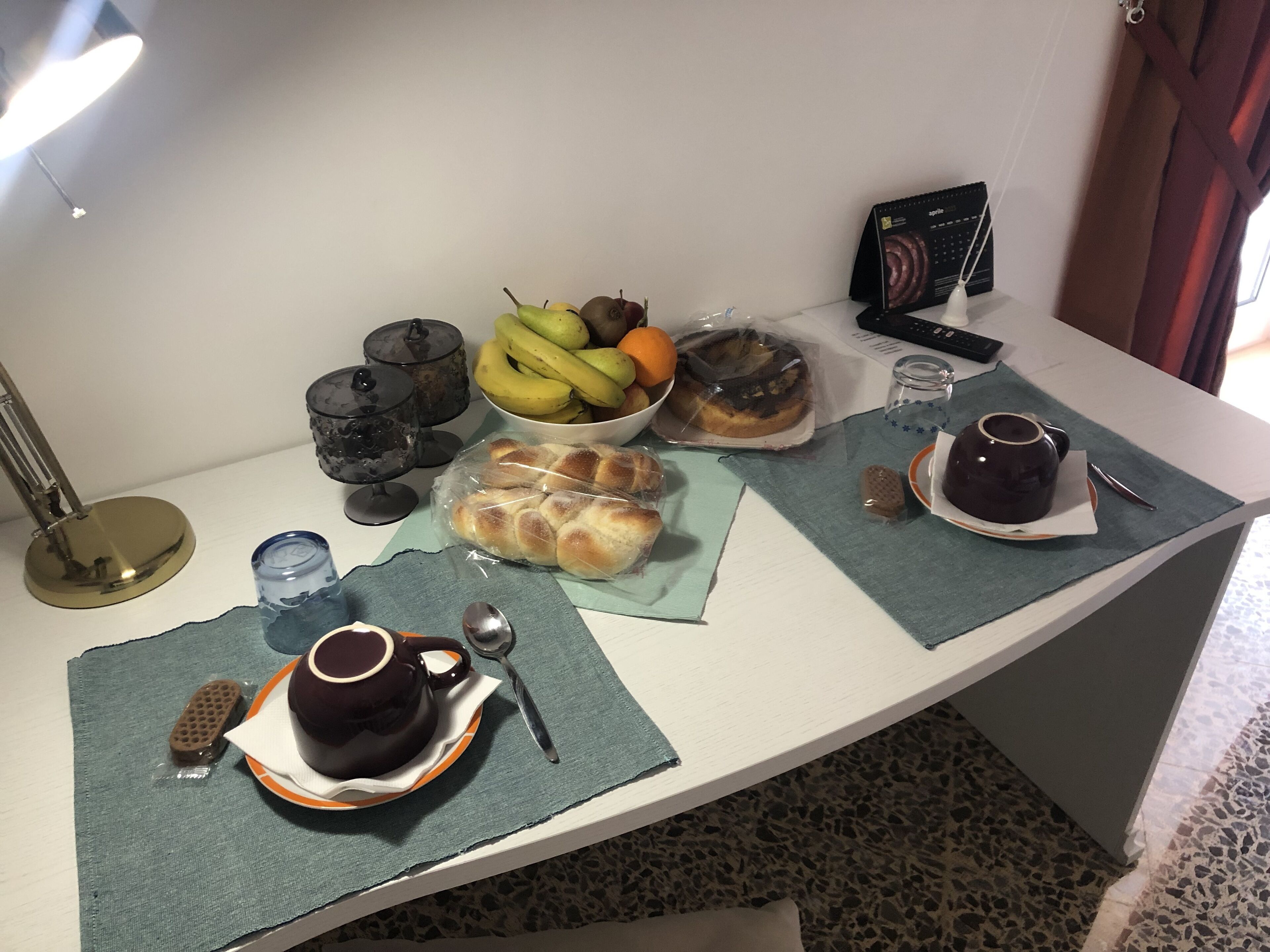 A breakfast setup with bread, fruits, a coffee pot, and a remote control on a table.