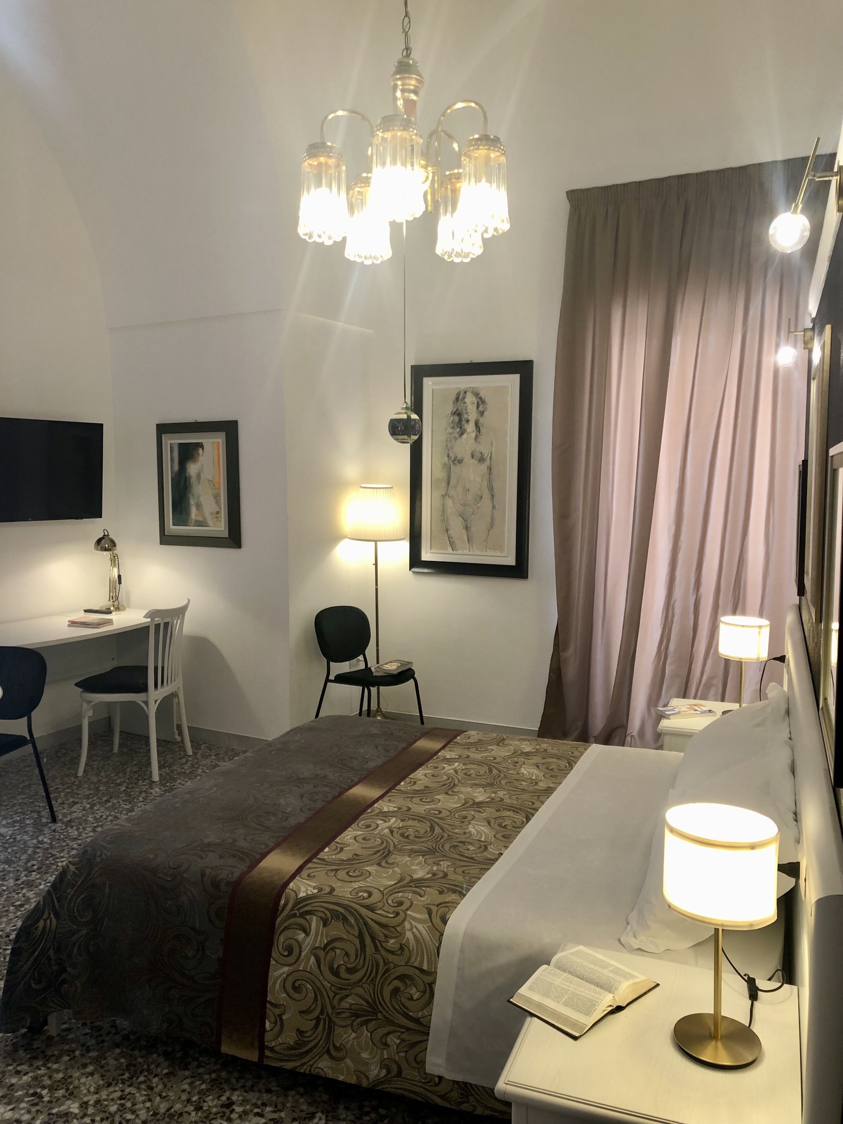 A hotel room with a bed, bedside table, lamp, chair, desk, and artwork on the wall.