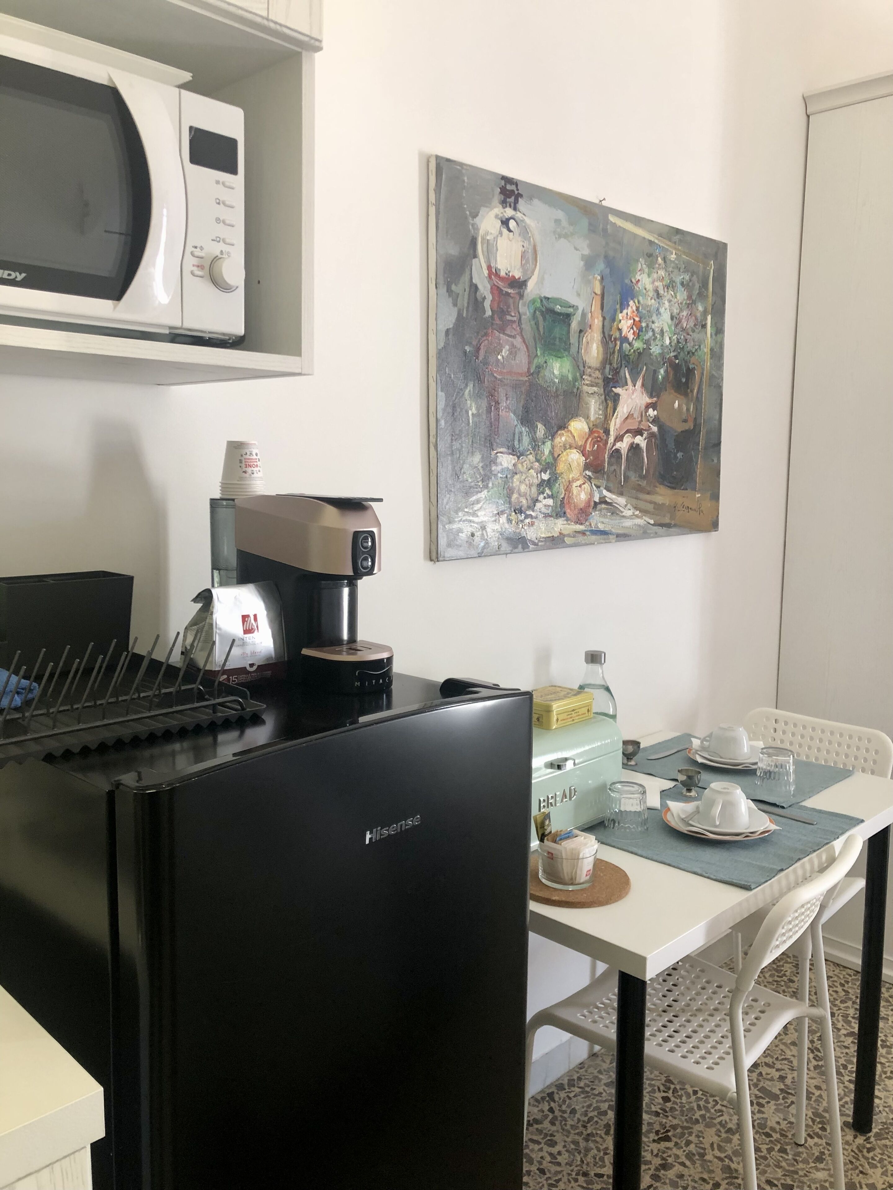 A compact kitchen with a microwave, coffee maker, and a Hisense refrigerator. There's a dining table with chairs and a painting on the wall.