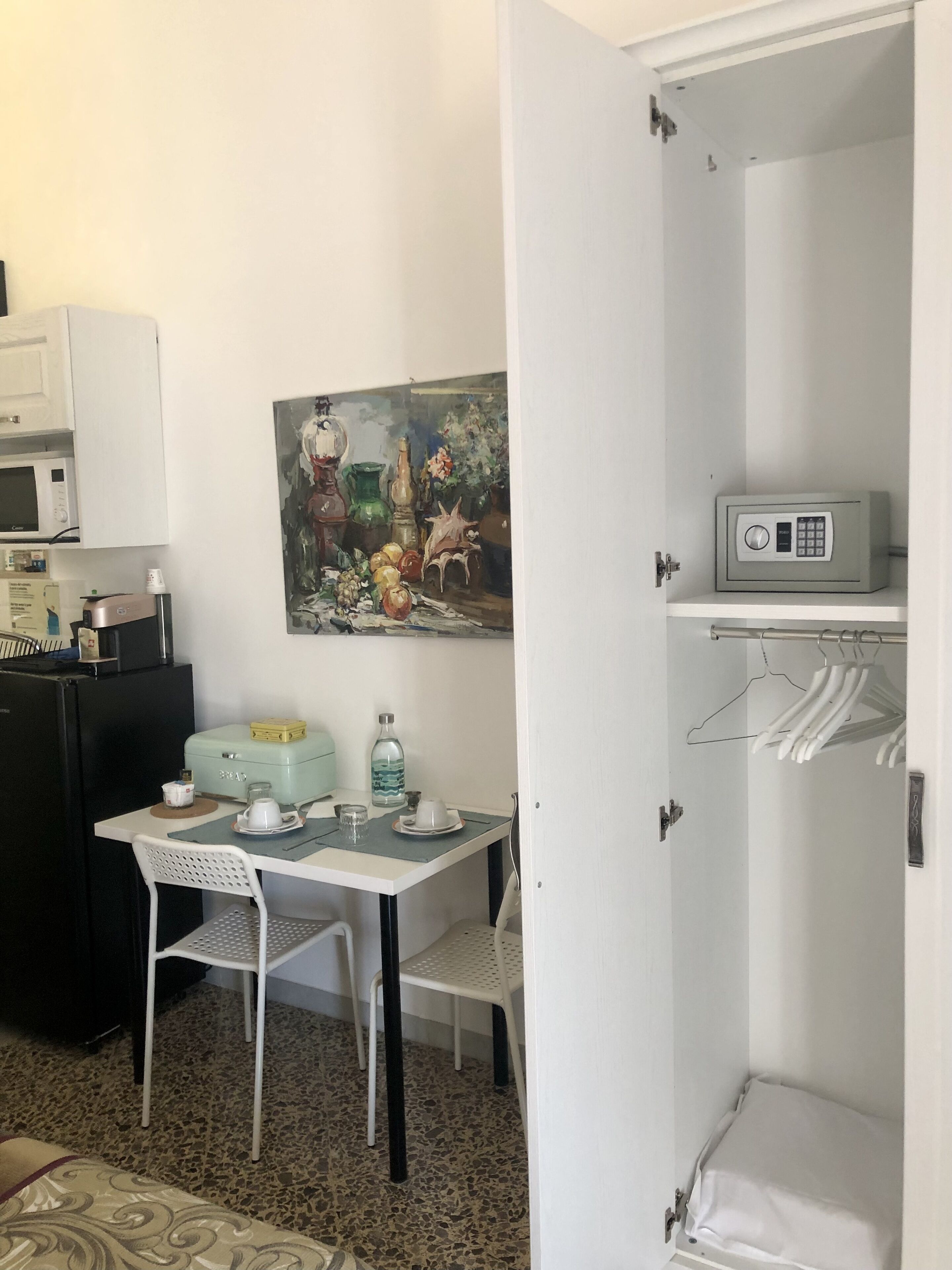 A compact room with a kitchenette, a dining table, a chair, a painting, and a wardrobe with hanging clothes.