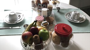 Et morgenbord med et udvalg af mad, blandt andet frugt, bagværk og tilbehør, placeret på et hvidt bord med udsigt til et vindue og gardiner.
