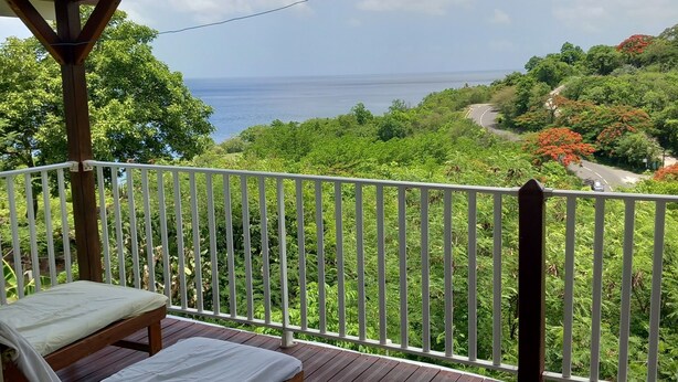 Eine Holzterrasse mit weißer Brüstung, einer Bank und Blick auf das Meer und grüne Vegetation.