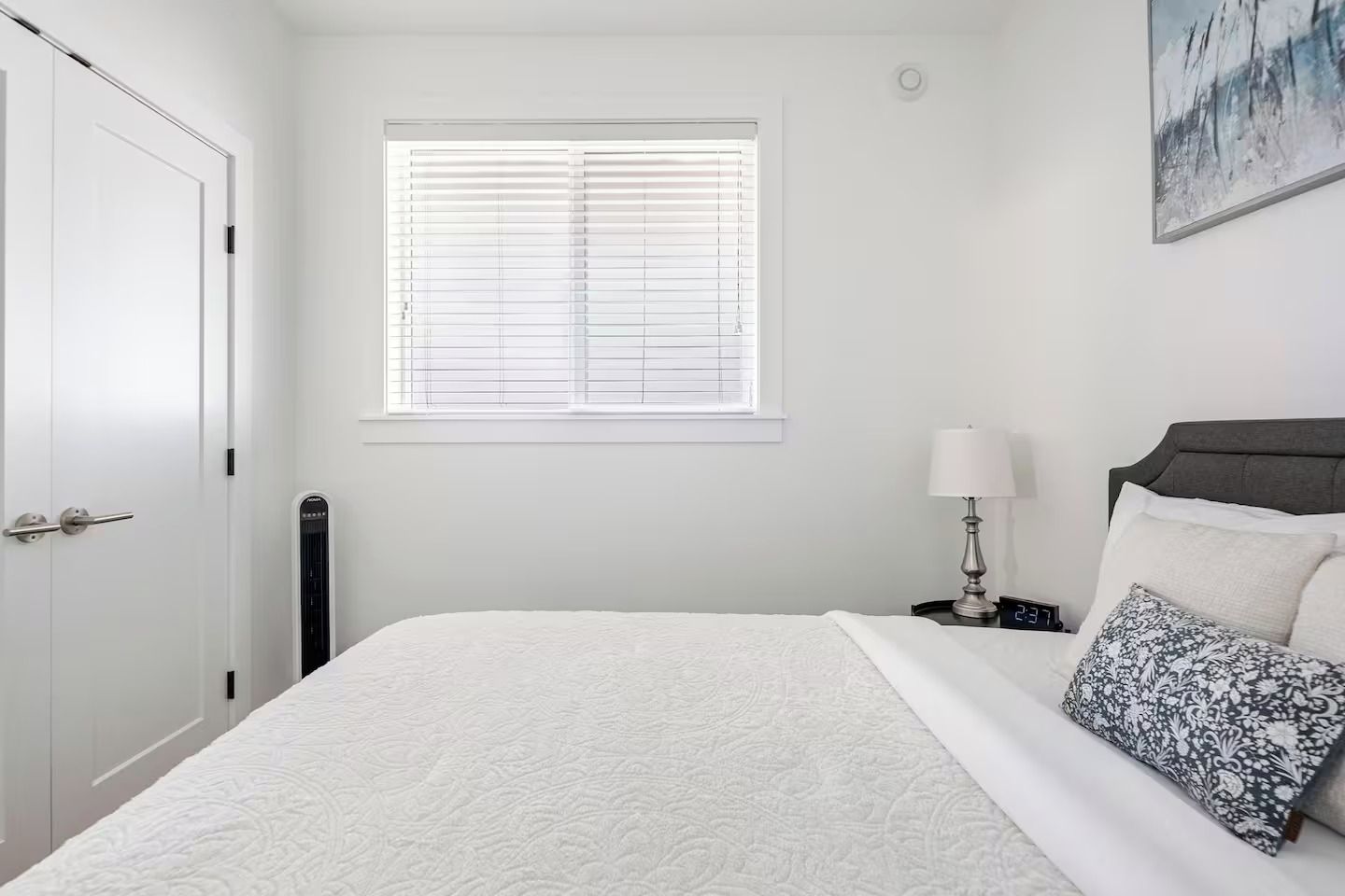 Un dormitorio con cama, mesita de noche, lámpara, reloj de pared y ventana con persianas.