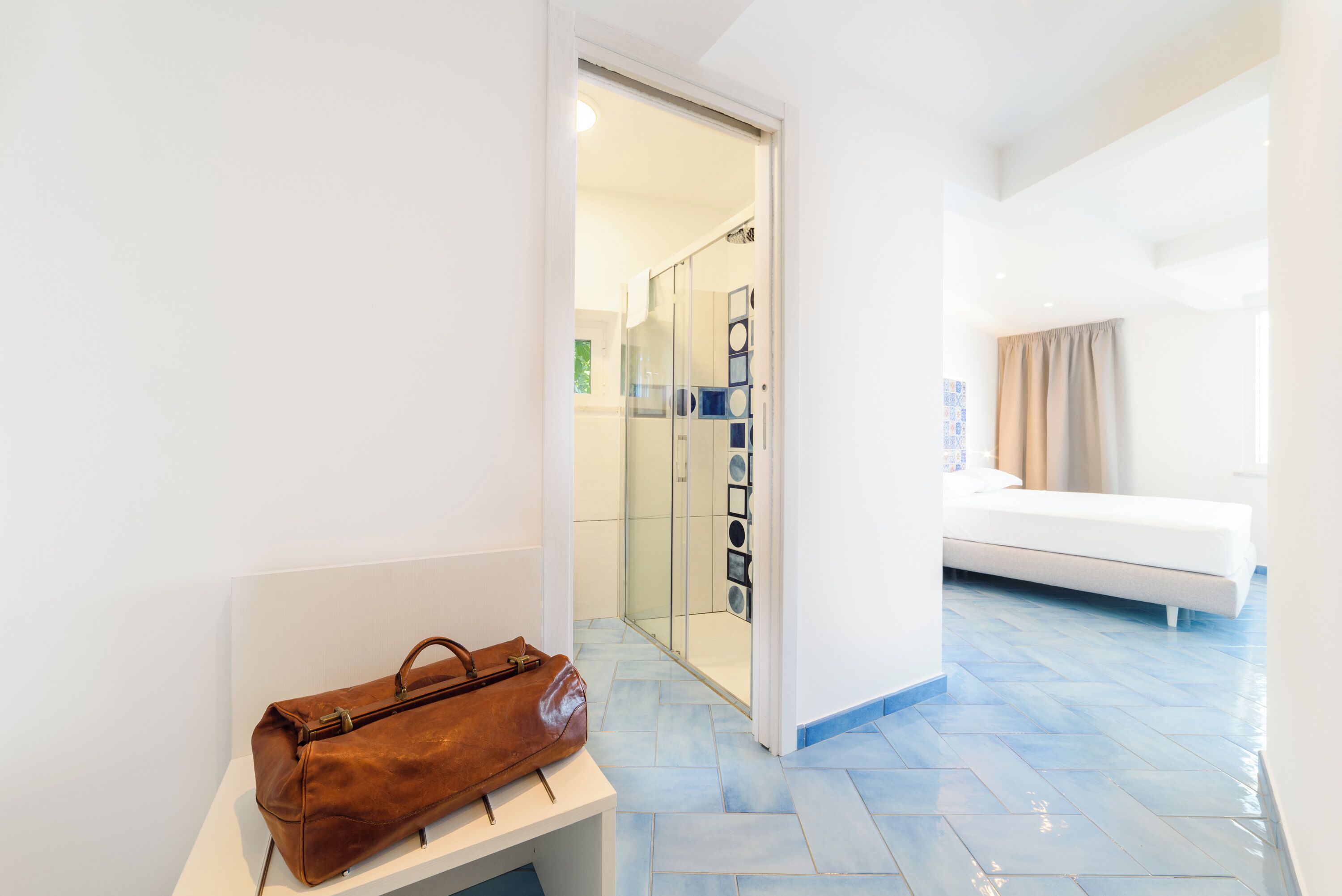 A hotel room with a bed, a bathroom with a glass shower, and a suitcase on a bench.