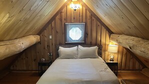 Accogliente camera mansardata con tetto spiovente in legno, un letto grande con biancheria bianca, un comodino con una lampada e una finestra con persiane.