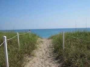 Un sentier sablonneux menant à la plage, bordé de gazon et délimité par des cordes blanches.