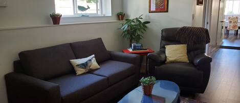 A living room with a brown sofa, a glass coffee table, a red side table, a black armchair, and a patterned rug.