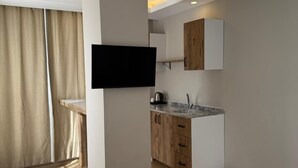 Una habitación de hotel moderna con sofá, una mesita redonda pequeña, televisor montado en la pared, kitchenette con armarios de madera y un ventanal con cortinas.