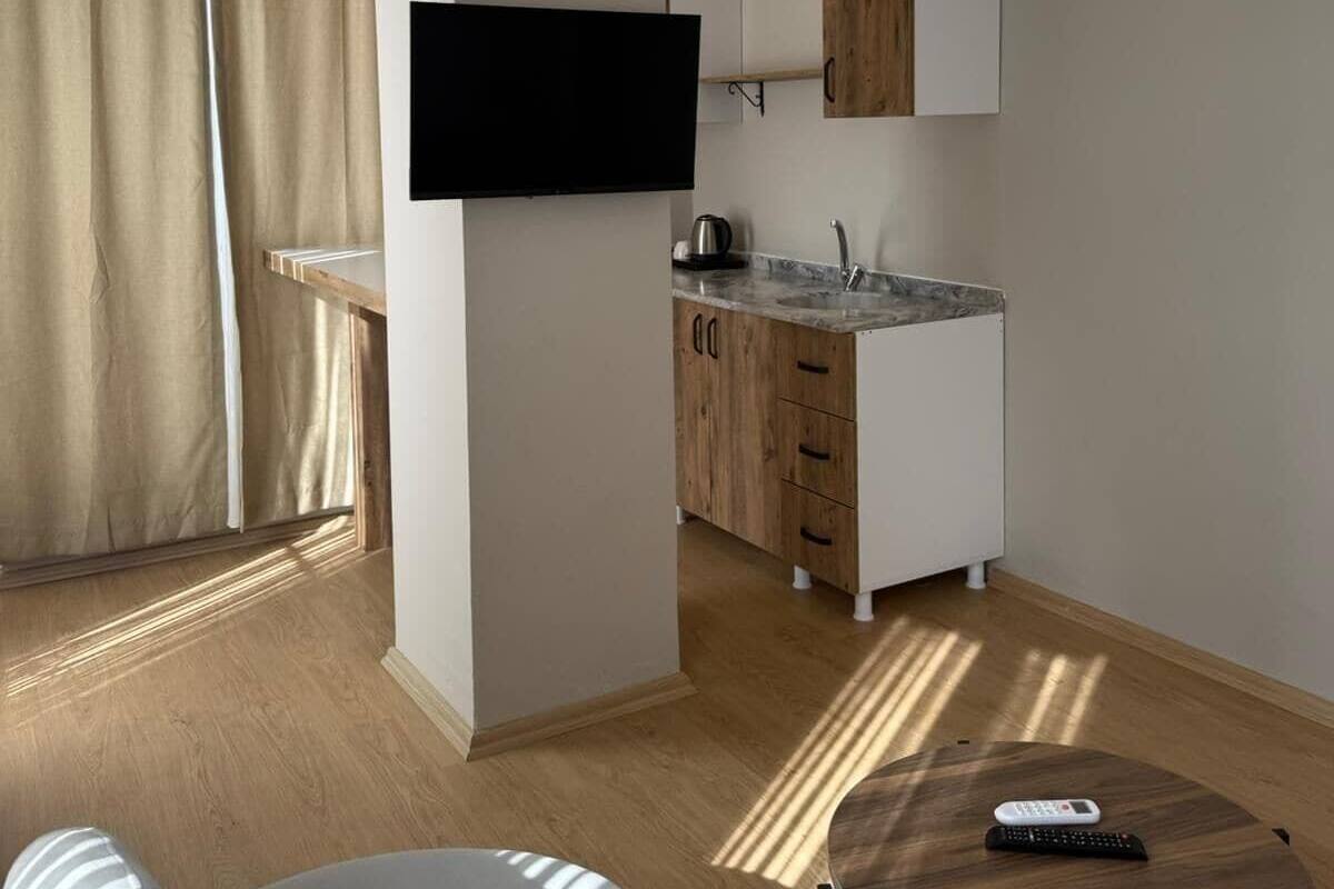 A modern hotel room with a sofa, a small round table, a TV mounted on a wall, a kitchenette with wooden cabinets, and a large window with curtains.