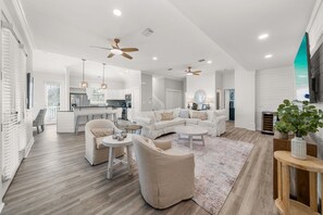 A spacious living area with a white sofa set, a coffee table, and a dining table. There is a kitchen in the background with modern appliances.