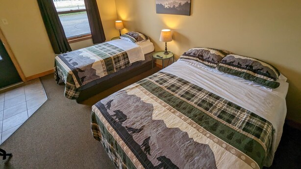 A room with two beds, each with a quilted bedspread featuring a mountain and forest design. There is a painting of a mountain landscape on the wall, and a window with curtains on the left side. - Chip-Wa Lodging Room 5 (Hunter)