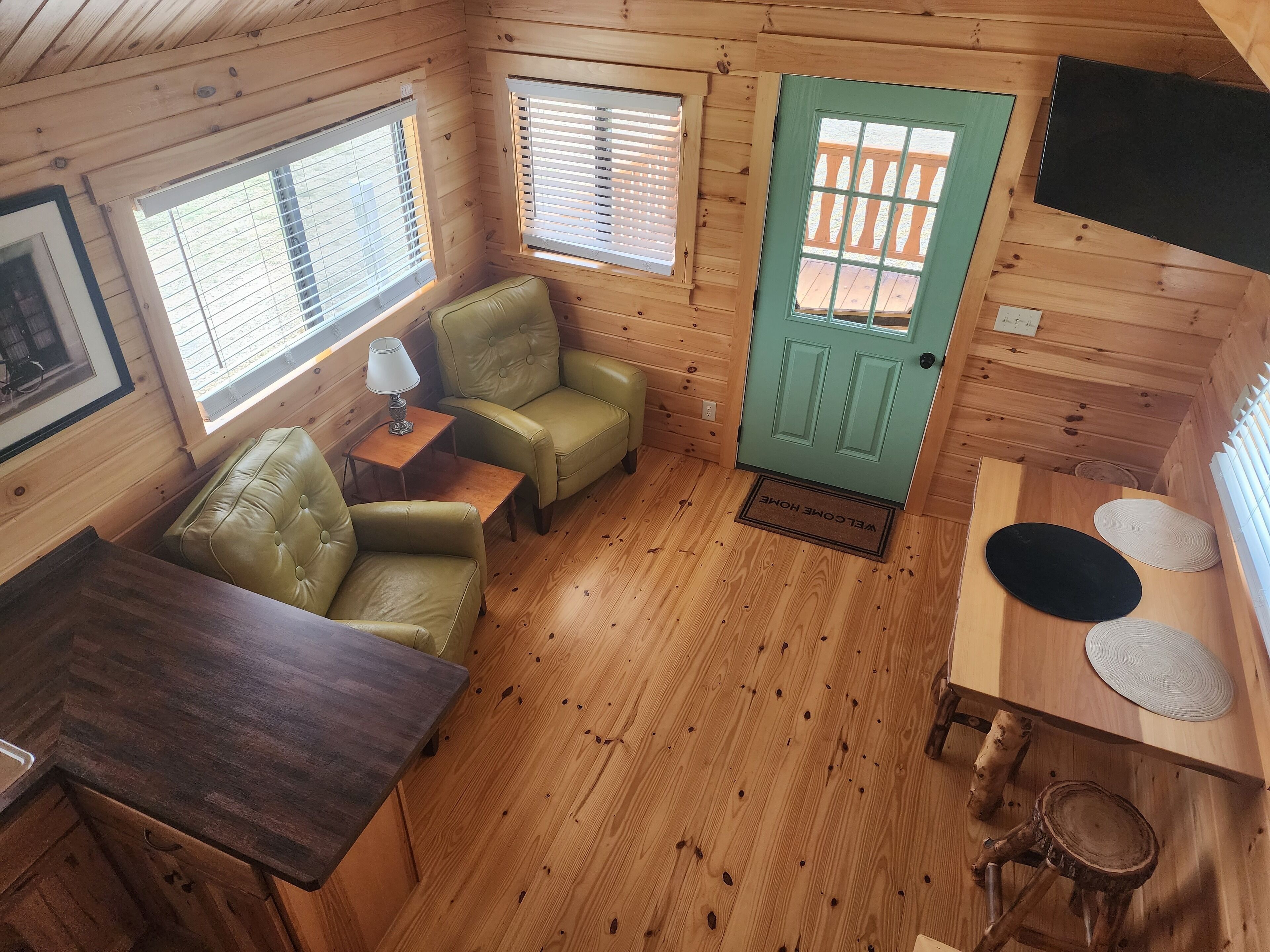A cozy cabin interior with a wooden floor, a green door, two windows with blinds, a small table with a lamp, and a green leather sofa.