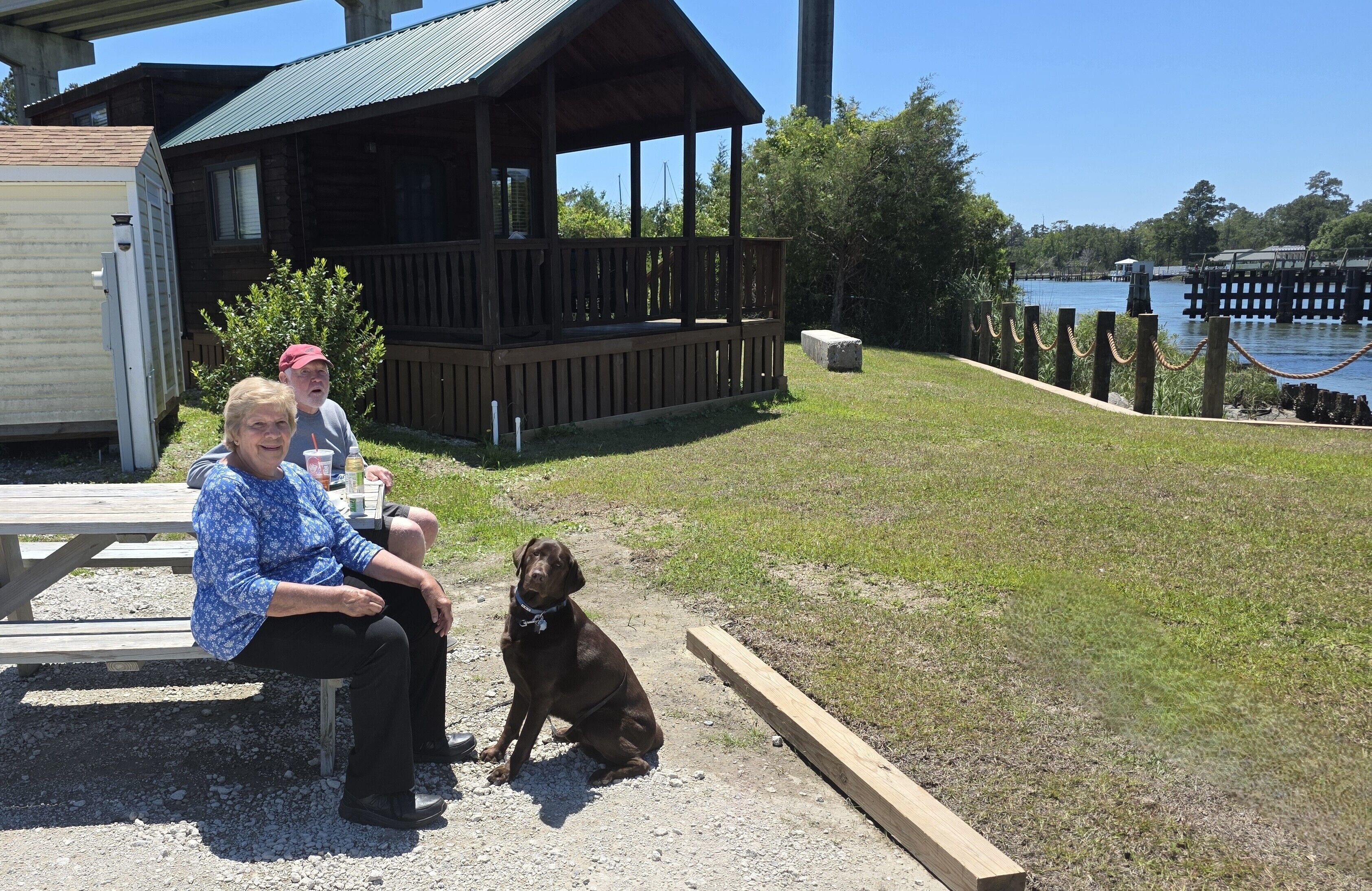 Una terraza de madera, una mesa de picnic, un perro y vistas al lago.