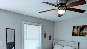 A bedroom with a bed, a ceiling fan, a window with blinds, a bedside table, and a wall-mounted mirror. - Perfectly Located Studio in MB (Myrtle Beach)