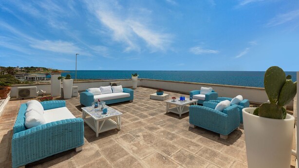A rooftop terrace with blue wicker furniture, a cactus plant, and a view of the sea.