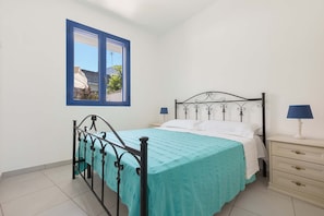 Una cama individual con estructura metálica, una ventana azul y una mesita de noche con lámpara.