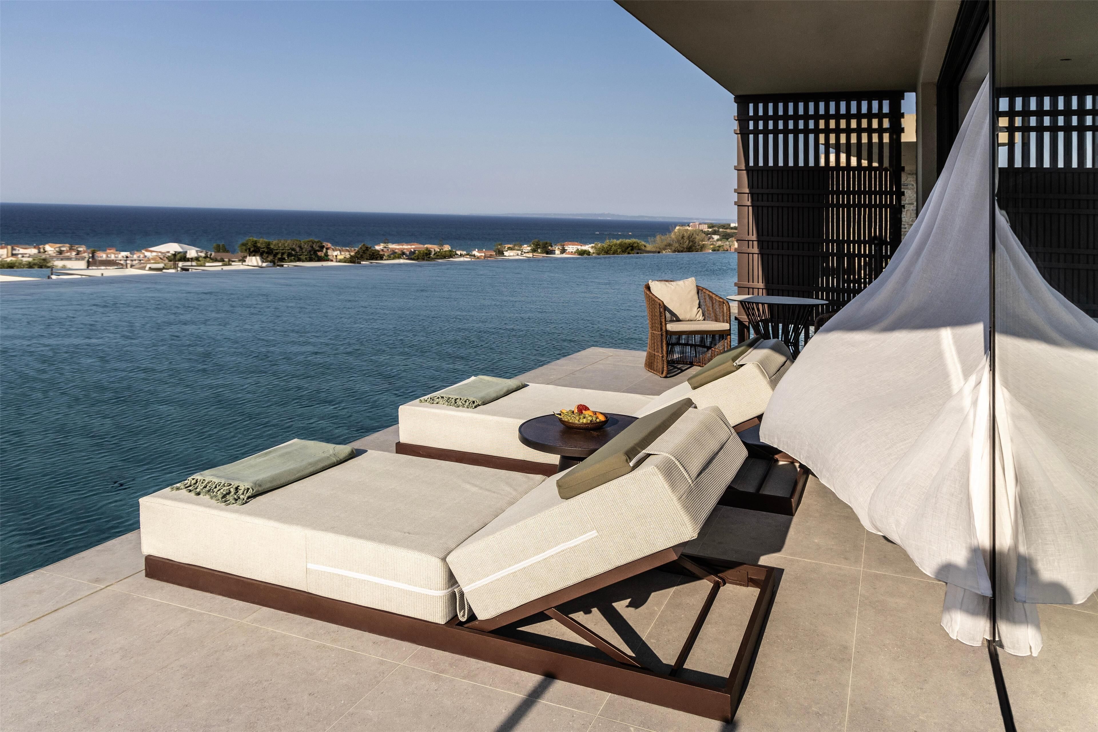 Un balcón con vista al mar, una silla de lounge, una mesa con un frutero y un dosel blanco.
