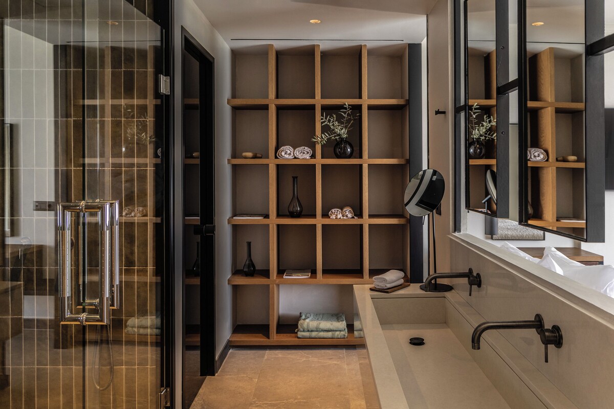 A modern bathroom with a glass shower enclosure, a built-in shelf unit with decorative items, and a large mirror above the sink.
