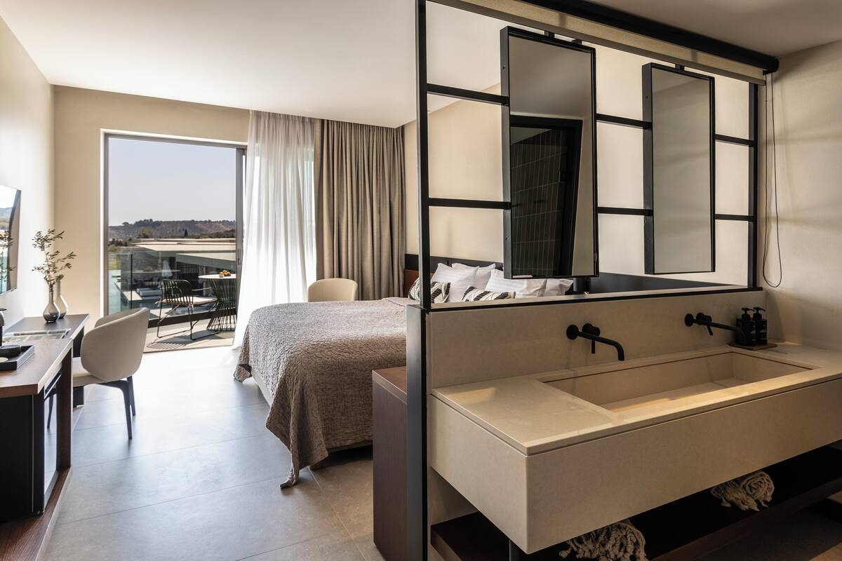 A modern hotel room with a large bed, a desk, a chair, a large window with a view, and a large bathroom with a double sink and a large mirror.