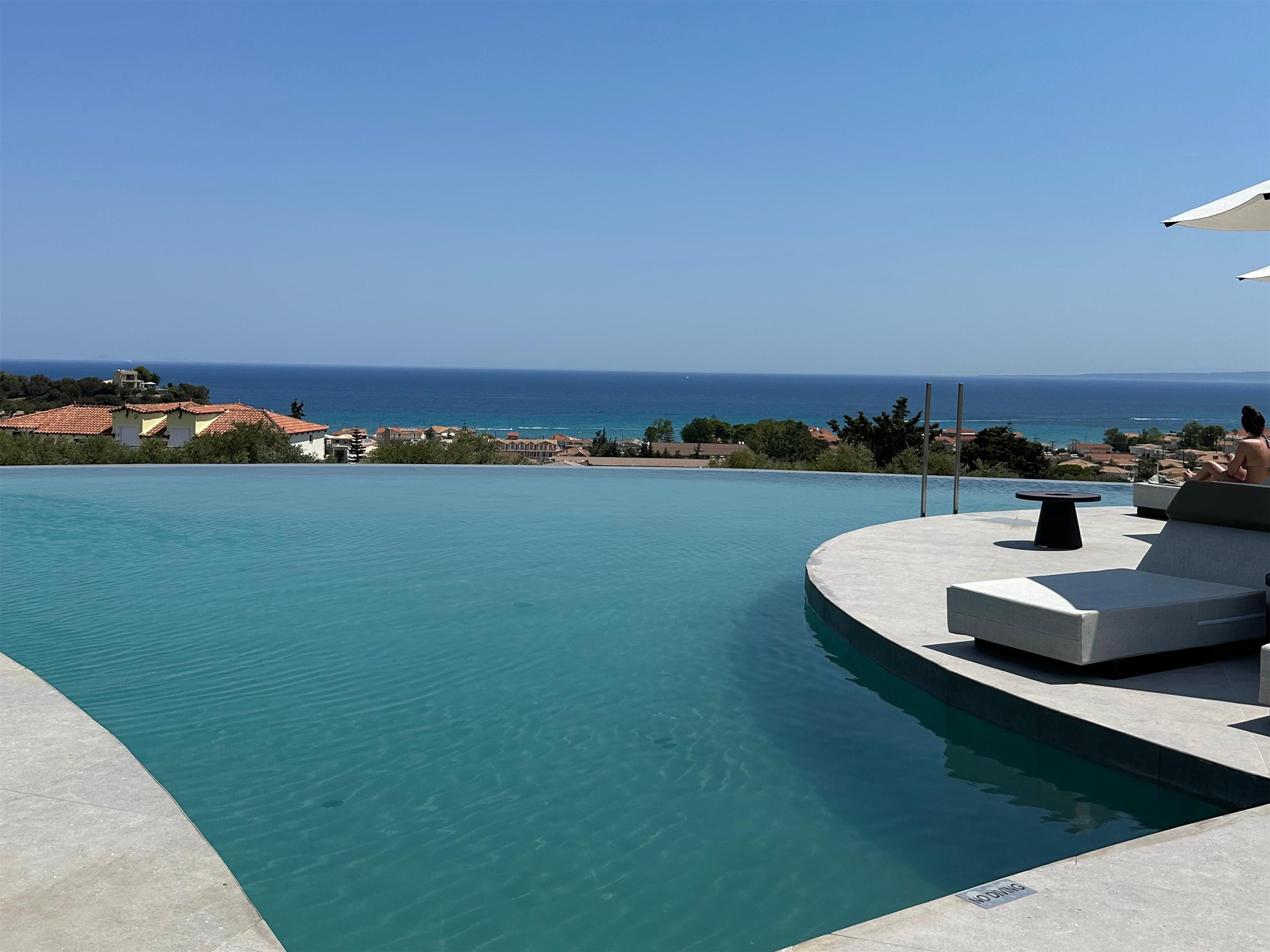 A swimming pool with a curved edge, a lounge area with a table, and a view of the sea and buildings in the background.