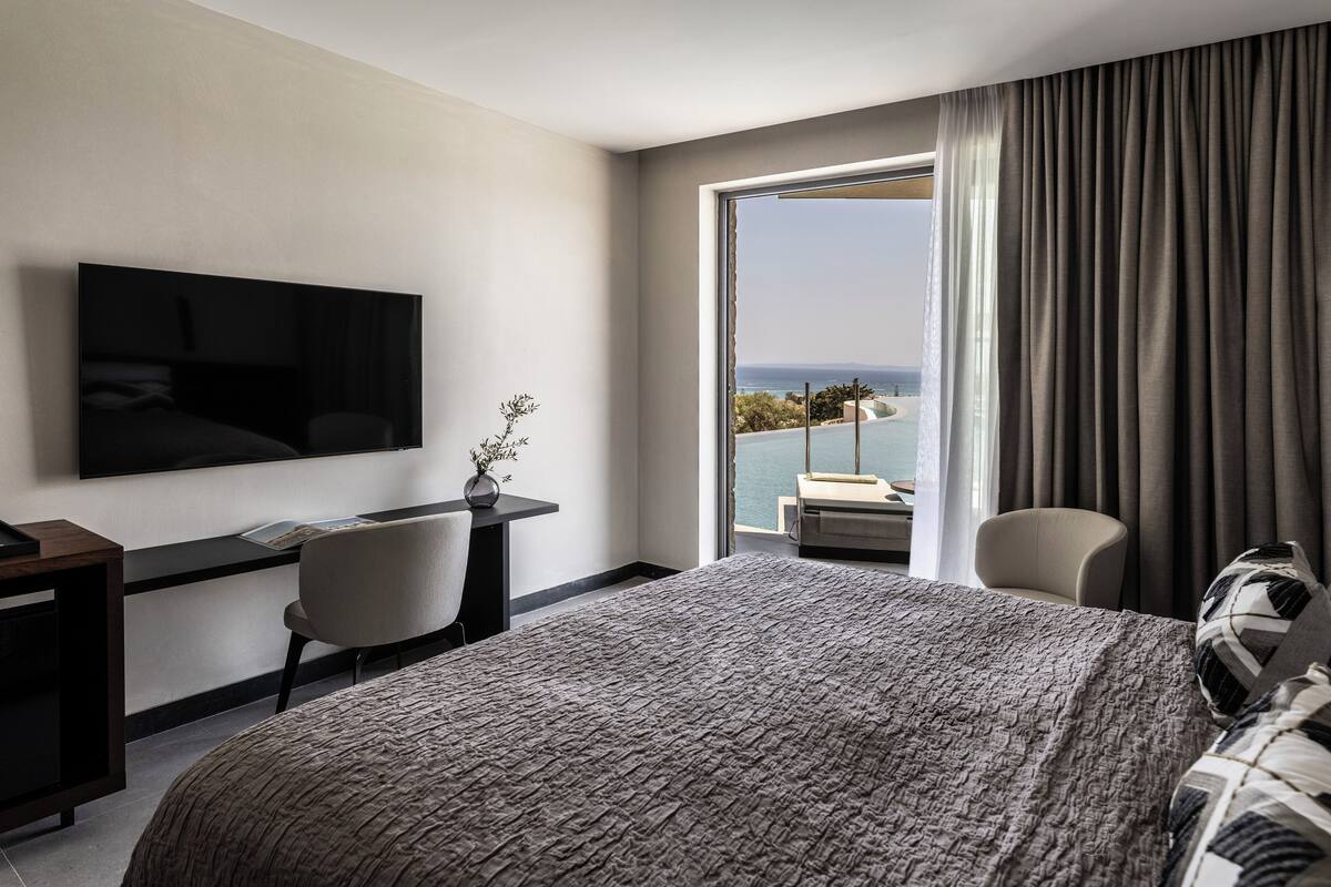 A hotel room with a large bed, a desk with a chair, a television mounted on the wall, and a balcony with a view of the sea.